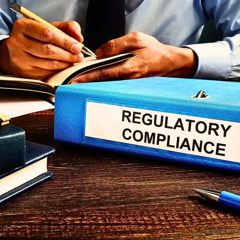 detecting-compliance-violations-blockchain-data Person writing in a notebook beside a binder labeled "Regulatory Compliance," symbolizing documentation and legal oversight
