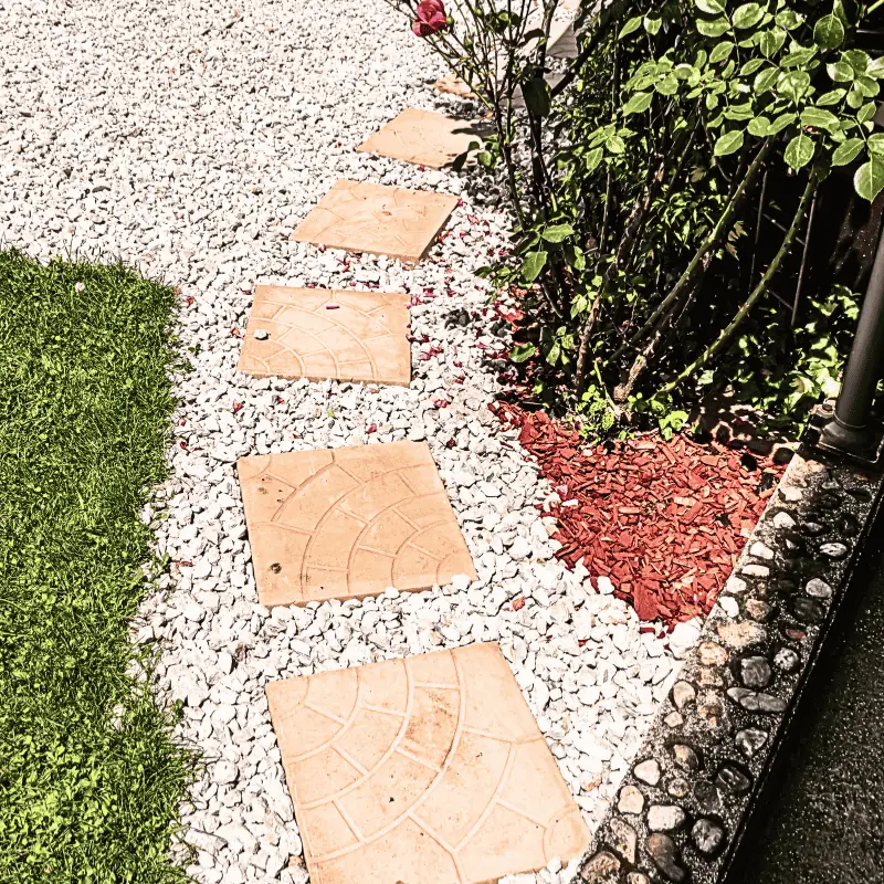 Square paver stones set in white gravel along a small garden edge with mulch and plants—an example of a low-maintenance, space-saving garden path.